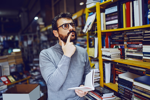 Attractive caucasian serious bearded man with eyeglasses standing in bookstore with book in hands and thinking.