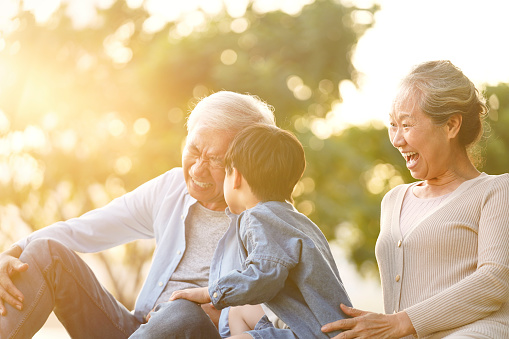 asian grandson, grandfather and grandmother sitting on grass having fun outdoors in park at sunset