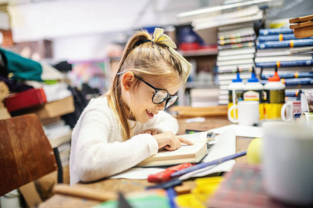 menina loura caucasiana adorável com rabo de cavalo e com os óculos que sentam-se na livraria e que lêem o livro emocionante. - picture book library preschool bookshelf - fotografias e filmes do acervo