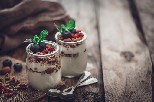 Healthy eating: two glass containers with homemade yogurt and granola shot on rustic wooden table. The composition is at the left of an horizontal frame leaving useful copy space for text and/or logo at the right. Selective focus on foreground. Retro style processed. Predominant color is brown. High resolution 42Mp studio digital capture taken with Sony A7rii and Sony FE 90mm f2.8 macro G OSS lens