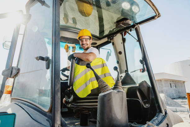 Smiling handsome caucasian worker in overall and with helmet on head driving excavator. Smiling handsome caucasian worker in overall and with helmet on head driving excavator. bulldozer photos stock pictures, royalty-free photos & images