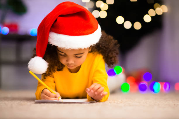 サンタ帽子書きのかわいい黒い女の子は、両親のためのリストをプレゼント書きます - wish list ストックフォトと画像
