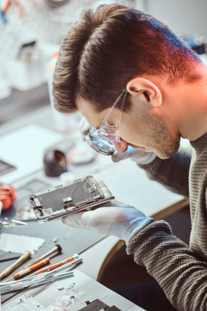 le technicien utilise une loupe pour inspecter soigneusement les parties internes du smartphone dans un atelier de réparation moderne - male magnifying glass caucasian technician photos et images de collection