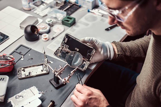 serviceman utilise un loupe et une pince à épiler pour réparer le smartphone endommagé dans l'atelier. - male magnifying glass caucasian technician photos et images de collection