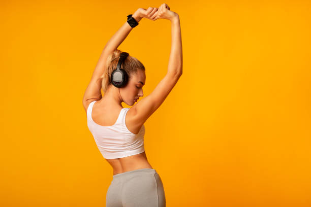 Unrecognizable Woman In Headphones Doing Stretching Exercises, Yellow Background Unrecognizable Woman In Headphones Doing Stretching Fitness Exercises Standing With Her Back To Camera Over Yellow Background. Copy Space blonde female bodybuilders stock pictures, royalty-free photos & images