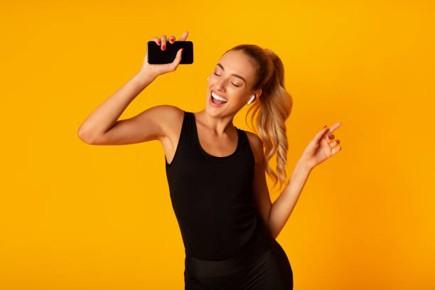 Woman In Wireless Earbuds Holding Smartphone And Dancing, Studio Shot Music App. Sporty Woman In Wireless Earbuds Holding Smartphone And Dancing Over Yellow Background. Studio Shot in ear headphones stock pictures, royalty-free photos & images