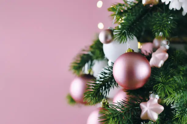 Photo of Close up of decorated Christmas tree with white seasonal and pink tree ornaments like baubles and stars on pink background with lights in background