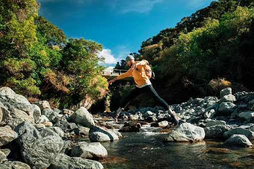A side-view shot of a young man crossing a stream of water, he is jumping from rock to rock.