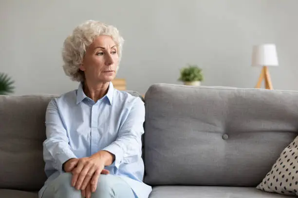 Thoughtful pensive senior grandma looking away thinking of loneliness sit on couch, melancholic worried serious old elderly woman lost in thought about depression wait support alone on sofa at home