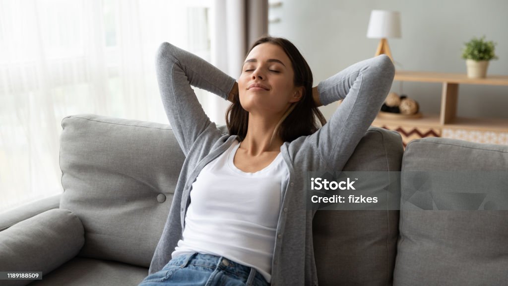 Relaxed serene young woman lounge on comfortable sofa at home Relaxed serene pretty young woman feel fatigue lounge on comfortable sofa hands behind head rest at home, happy calm lady dream enjoy wellbeing breathing fresh air in cozy home modern living room Relaxation Stock Photo