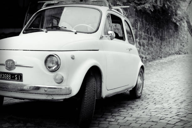 Cremona, Italy June 11th 2025 Low angle view of a vintage red Fiat 500 parked on an asphalt road, surrounded by modern cars, residential buildings, and trees, capturing Italian urban nostalgia