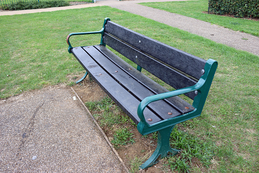 Green metal framed recycled black plastic seat in a park with a grass lawn with tarmac paths in the background.