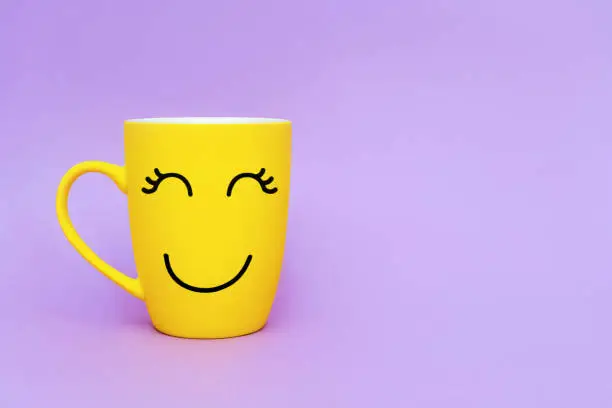 Photo of Smiley yellow coffee cup on purple background. Happy friday word concept. Minimalism style, romantic mood, good morning, happiness, break time