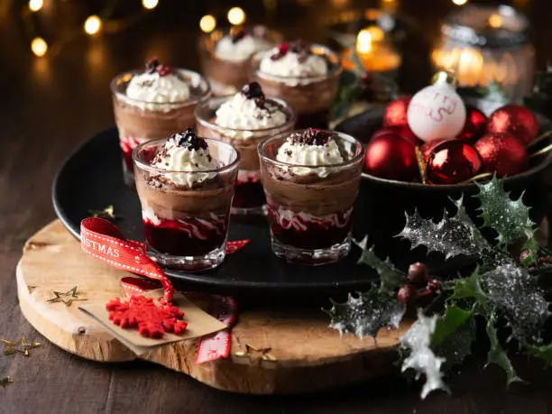 Photo of Dessert in a glasses with chocolate and berries spread on wooden background with garland lights bokeh and christmas decoration. New year holidays background concept. Dessert recipe ideas.