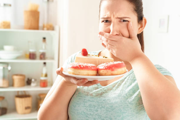 istock Fighting Temptation. Chubby girl standing in kitchen holding plate with desserts scared close-up 1189231183