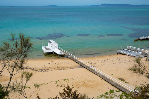 Long wooden pier extending over the beach into shallow turquoise bay water with white wooden shed at the tip