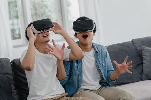 Young Asian Gay couple using technology funny at home, Asia lover guy lgbtq+ feeling happy fun and virtual reality, VR playing games together while lying sofa in living room at home concept.