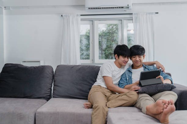 junge homosexuell paar mit tablet zu hause. asiatische lgbtq+ männer glücklich entspannen spaß mit technologie beobachten film im internet zusammen beim liegen sofa im wohnzimmer konzept. - pride lgbtqi veranstaltung stock-fotos und bilder