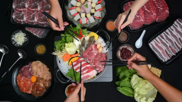 Top view of people eating Shabu-Shabu in hot pot with fresh sliced meat, sea food, and vegetables with black background, Japanese cuisine