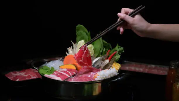 Cropped shot of woman eating Shabu-Shabu in hot pot with fresh sliced meat, sea food, and vegetables with black background, Japanese cuisine