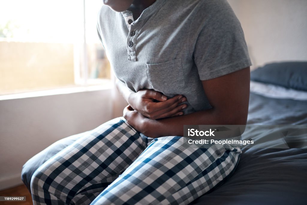 Ouch! My tummy! Cropped shot of an unrecognizable man sitting alone on his bed and suffering from stomach cramps while home alone Stomachache Stock Photo