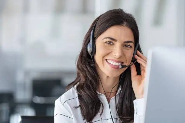 Call center agent with headset working on support hotline in modern office with copy space. Portrait of mature positive agent in conversation with customer over headset looking at camera. Consulting and assistance service customer support.