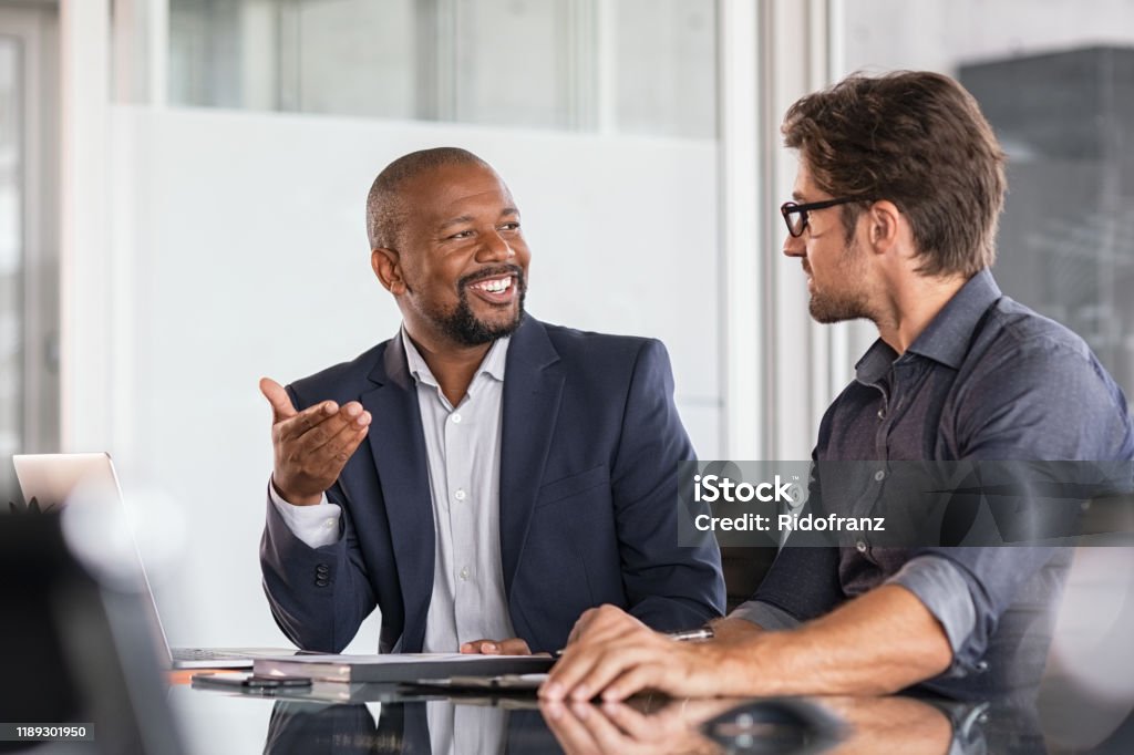Multiethnic business people in meeting Two happy business colleagues at meeting in modern office interior. Successful african boss in a conversation with young employee in boardroom. Marketing team of two businessmen discussing strategy in meeting room. Discussion Stock Photo