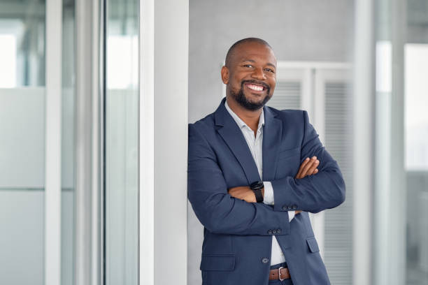 Successful businessman in modern office Mature cheerful african american executive businessman at workspace office. Portrait of smiling ceo at modern office workplace in suit looking at camera. Happy leader standing in front of company building. mature men photos stock pictures, royalty-free photos & images