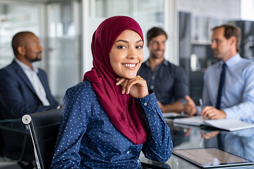 Beautiful arab businesswoman looking at camera and smiling while working in office. Portrait of cheerful islamic young woman wearing hijab at meeting. Muslim business woman working and sitting at conference table with multiethnic colleagues in background.