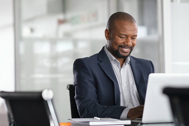 Black mature businessman working on laptop Positive mature businessman working on laptop in modern office. Successful african business man working on computer while sitting at desk. Smiling middle aged man working in a corporate. laptop businessman business men stock pictures, royalty-free photos & images