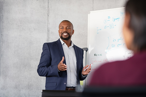 Mature african american coach explaining strategy to his team with copy space. Successful businessman presenting new project to employees during meeting room. Conference speaker talk to audience while giving presentation on whiteboard to business group of people.