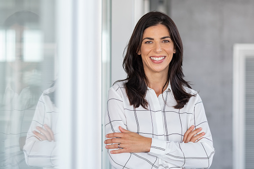 Portrait of middle aged businesswoman in modern office looking at camera. Confident business woman with arms crossed standing while leaning against glass wall. Proud brunette woman smiling in formalwear with copy space.