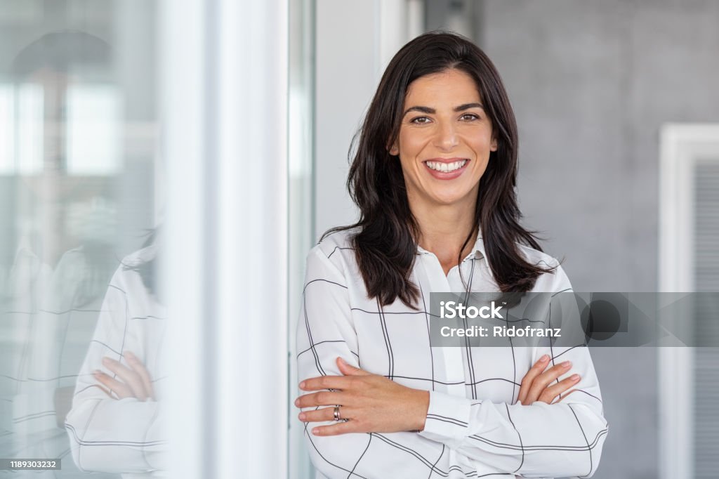 Successful mature business woman looking at camera Portrait of middle aged businesswoman in modern office looking at camera. Confident business woman with arms crossed standing while leaning against glass wall. Proud brunette woman smiling in formalwear with copy space. Women Stock Photo