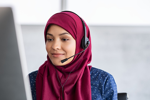 Muslim business woman with headset working in office. Happy arab woman working in company service center wearing headphone and using computer helping solving client problem. Islamic business woman working on computer at call center with copy space.