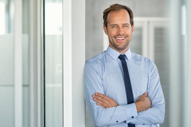 Successful mature business man in modern office Portrait of a successful leader smiling at modern office with copy space. Happy executive businessman looking at camera at work place. Mature entrepreneur standing with crossed arms leaning on glass wall with copy space. in pride we trust stock pictures, royalty-free photos & images