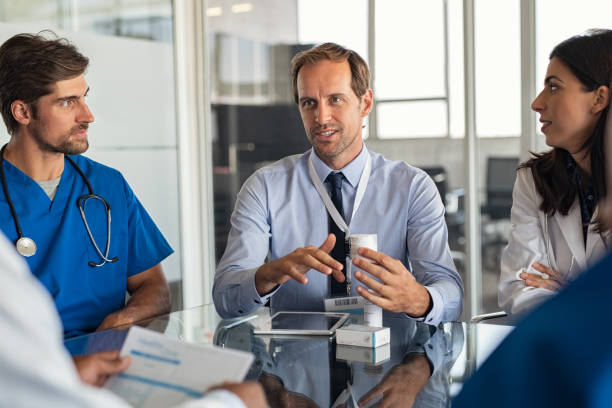 Pharmaceutical advisor showing new medicine Mature man with professional doctors in hospital showing medicine bottles. Confident representative pharmaceutical showing new medicines to a team of doctor and nurse. Specialist showing bottle of pills and explaning drug dosage to a group of specialist in meeting room. pharmaceutical industry stock pictures, royalty-free photos & images