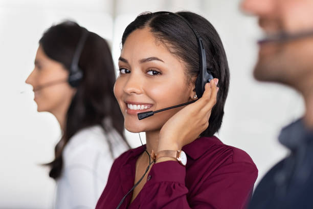 Young latin woman in customer service center Smiling beautiful woman working in call center as telemarketing operator. Smiling customer support agent wearing headset at office, working while looking at camera. Middle eastern girl working as telephone operator with her colleagues. people in a row photos stock pictures, royalty-free photos & images