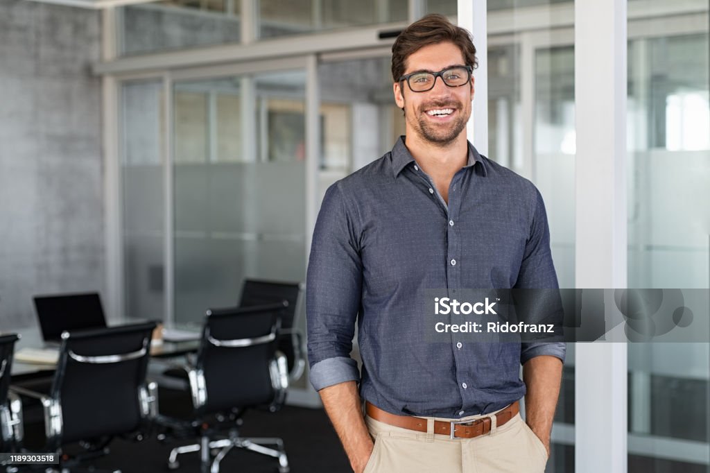 Success businessman smiling in office Portrait of young businessman wearing eyeglasses and standing outside conference room. Portrait of happy business man wearing spectacles and looking at camera with copy space. Satisfied proud man feeling confident in a modern office. Men Stock Photo