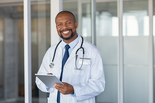 Portrait of confident mature black doctor consulting digital tablet and looking at camera. Smiling african american doctor with stethoscope using tablet at medical clinic. Happy healthcare worker using computer at modern hospital.