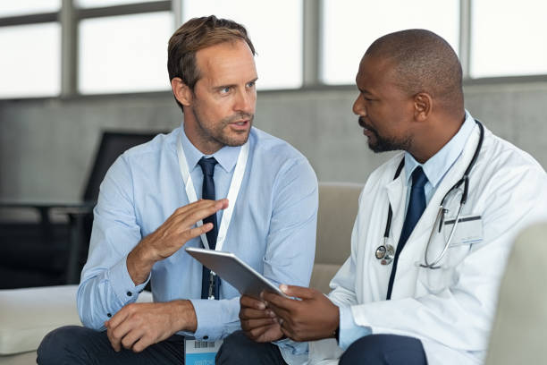 Doctor in conversation with pharmaceutical representative Pharmaceutical representative talking with doctor sitting on couch. African mature practitioner discussing results of the analysis with specialist while consulting diagnosis on digital tablet. Doctor in conversation with medical advisor in hospital room discussing patients report case. medical occupation stock pictures, royalty-free photos & images