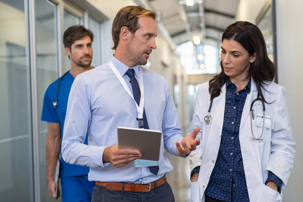 Doctor in a conversation with specialist Man and woman doctor having a discussion in hospital hallway while holding digital tablet. Doctor discussing patient case status with his medical staff after operation. Pharmaceutical representative showing medical report. doctor consultation stock pictures, royalty-free photos & images