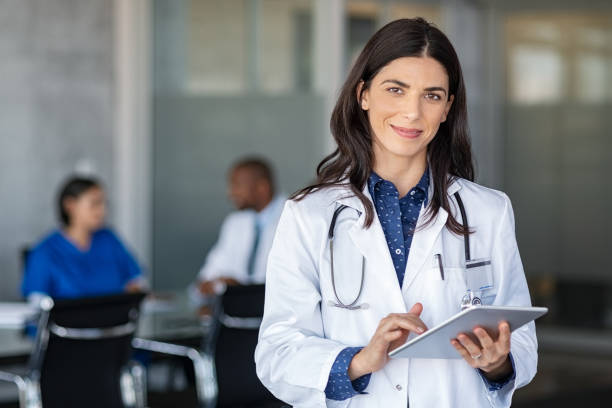 Doctor holding digital tablet at meeting room Portrait of beautiful mature woman doctor holding digital tablet and looking at camera. Confident female doctor using digital tablet with colleague talking in background at hospital. Latin nurse in labcoat and stethoscope in private clinic with medical team working. laboratory coat stock pictures, royalty-free photos & images