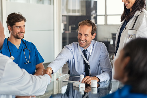 Happy businessman shaking hands with doctor in meeting room. Doctor and representative pharmaceutical shaking hands in medical office. Cheerful salesman with new medicines shaking hands with senior doctor in hospital with medical team sitting at conference table.