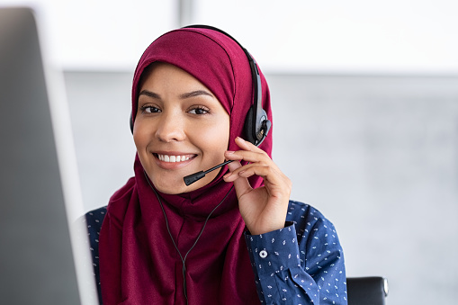 Portrait of happy young islamic woman in headscarf holding microphone and working on computer. Arab businesswoman working in customer service center talking using headset. Portrait of beautiful arabic woman in hijab smiling and looking at camera while working in office with copy space.