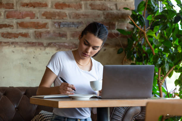 istock Young business woman sitting at table and taking notes in notebook.On table is laptop, smartphone and cup of coffee.On computer screen graphics and charts. Student learning online. Blogger. 1189305684