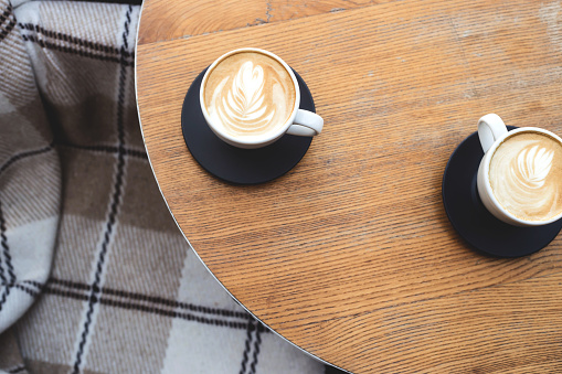 Two cups of cappuccino with latte art on a wooden table next to checkered cozy armchair . The concept of break in the city.