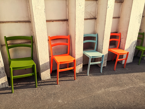 A row of multicolored plastic chairs stands along a white wall in the street, outdoors. Five stools (orange, green, blue) stand on the pavement, beautiful colorful background with copy space