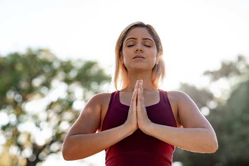 Young blonde woman standing with her hands together and her eyes closed in a park meditating on a sunny day