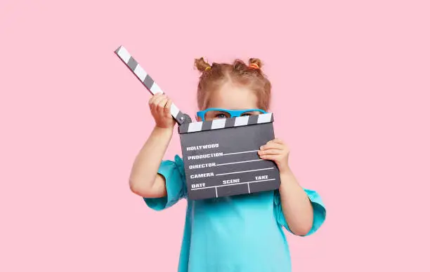 Photo of Funny smiling child girl in cinema glasses hold film making clapperboard isolated on pink background. Studio portrait. Childhood lifestyle concept. Copy space for text