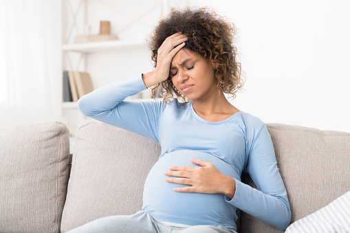 Pregnant afro girl suffering from headache and touching belly, sitting on sofa